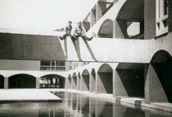 A black and white photograph of two young men sitting on a beam over the quad at the University of Sussex